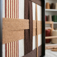 An angled close-up view of the framed fiber art in a studio setting, highlighting the three-dimensional depth of the woven soil-inspired yarn.