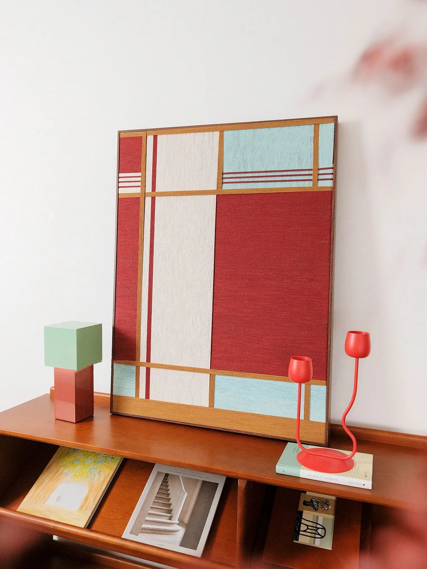 Red House yarn wall art styled on a desk beside stationery and a mint-green block ornament, showcasing its compact square format and vivid color blocking.
