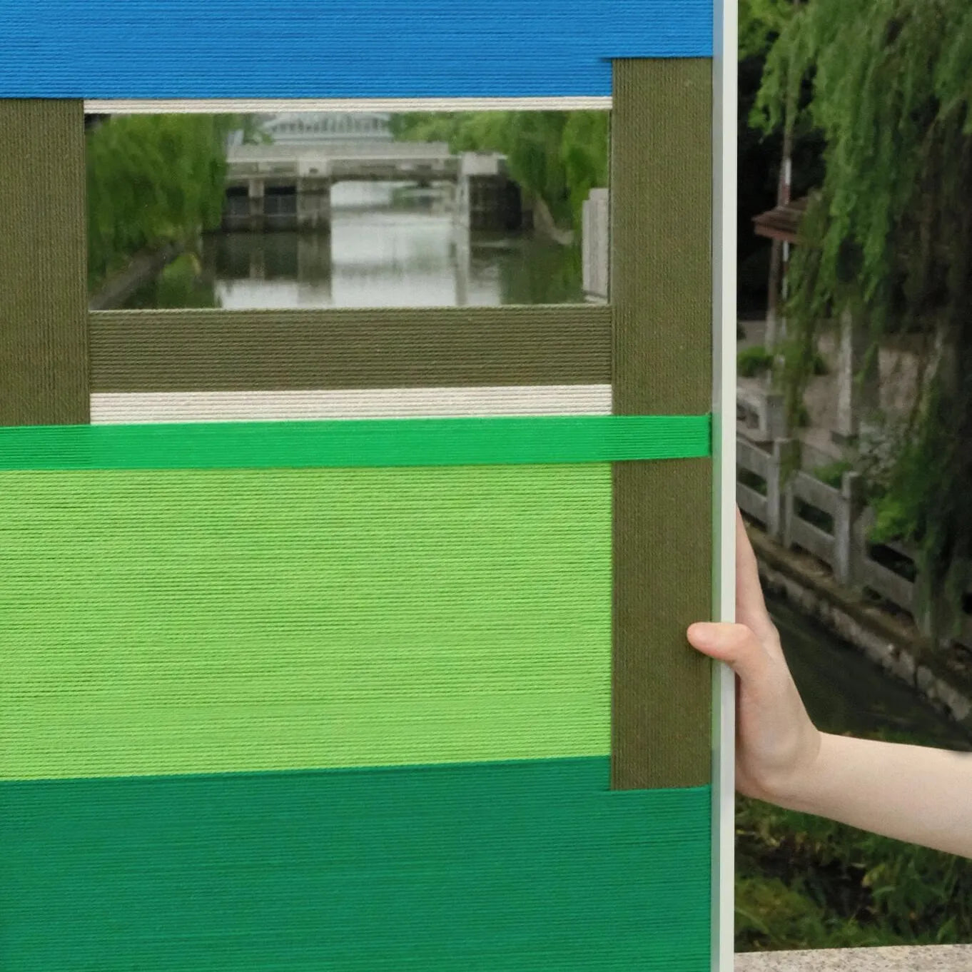 Hand-held view of Riverside yarn wall art showing its compact square proportions and natural wood frame with precise stripe alignment
