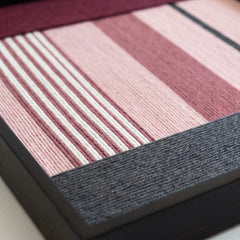 Angled close-up of the Rosy Pulse fiber art showing the dense hand-woven wool texture and sleek black metal frame finish.