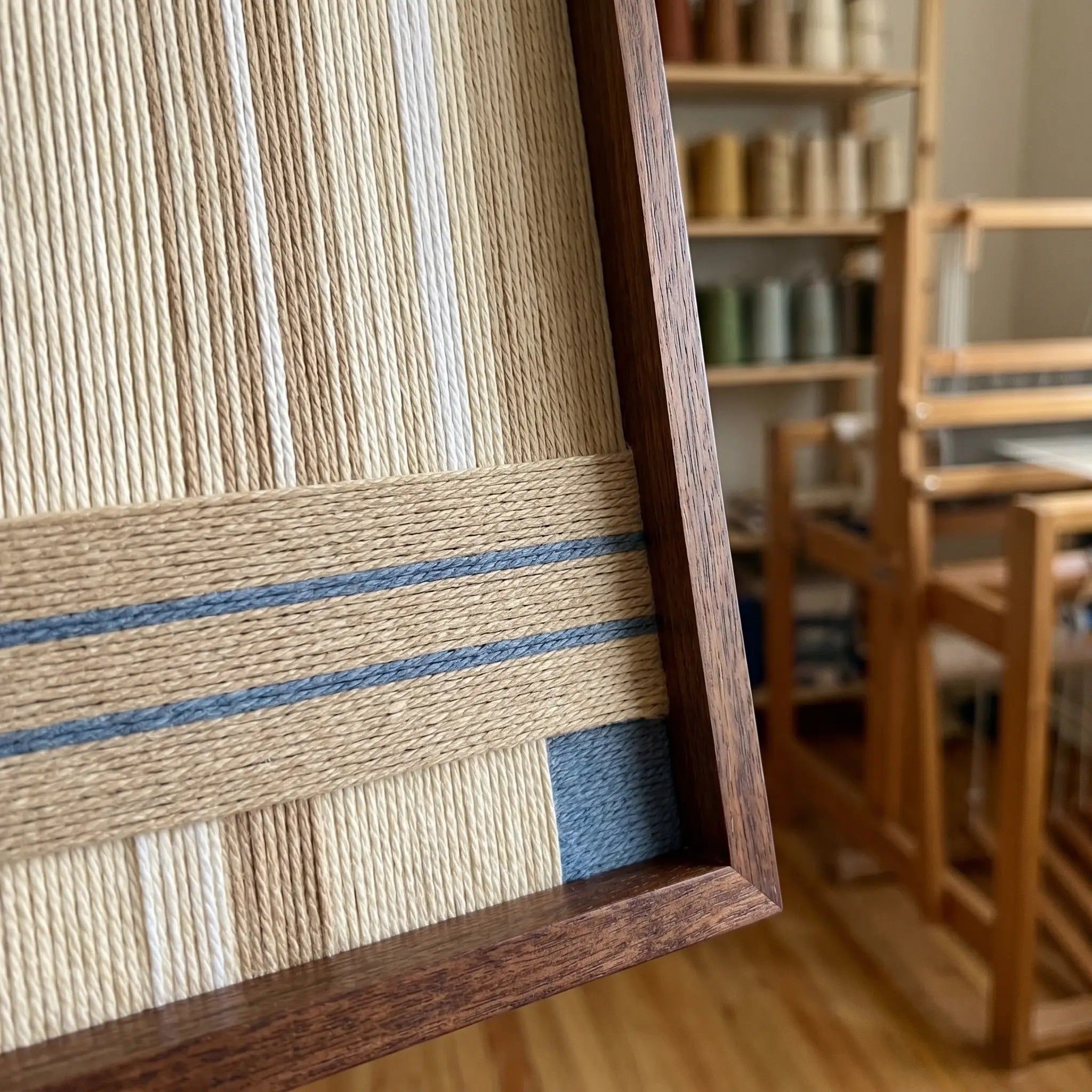 Hand-woven wool tapestry shown from an angled perspective in a sunlit artist studio with a wooden weaving loom in the background.