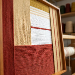 An angled close-up view of the Sun-Baked Terra wool art showing the vibrant terracotta and sand colored threads in a textured geometric layout.