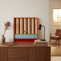 Square striped textile wall art hanging above a dark wood dresser or sideboard in a warmly lit room.