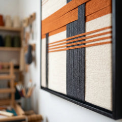 Angled side view of the framed fiber wall art hanging in a studio, showcasing the dimensional depth of the woven terracotta and grey yarn against the black frame.