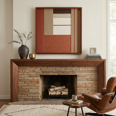Beautiful framed hand-woven textile wall art styled elegantly above a rustic stone and wood fireplace mantel in a cozy living room with a leather chair.