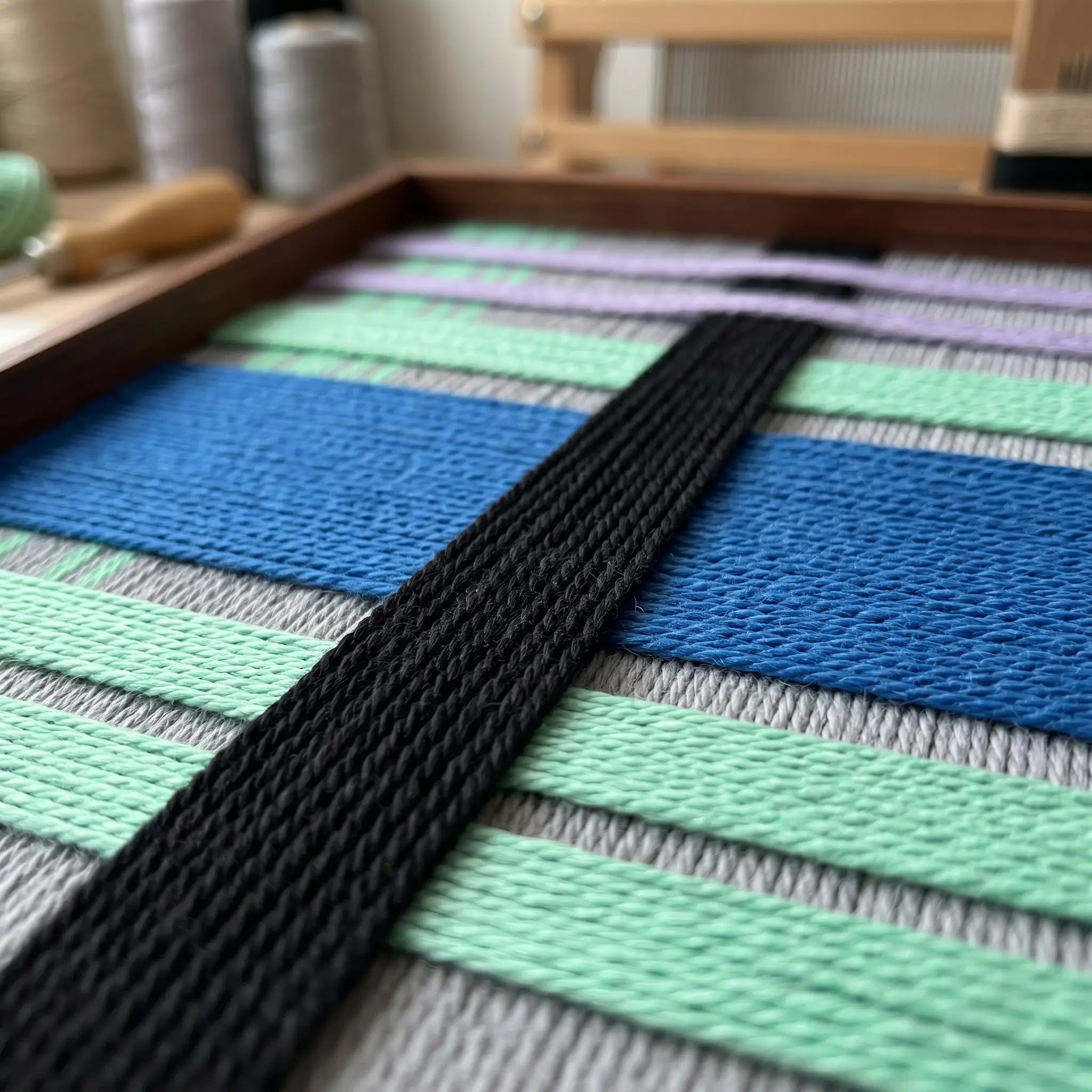 Angled macro shot of the Winter Aurora fiber art in a studio, highlighting the raised black vertical yarn woven over horizontal cobalt blue and mint green background layers.