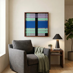 Square woven wool tapestry with vibrant blue and green geometric patterns displayed above a grey armchair in a cozy reading corner.