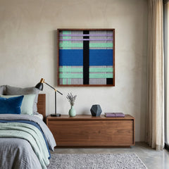 Framed colorful hand-woven wool wall art styled in a modern bedroom hanging above a wooden dresser next to a bed with matching blue tones.