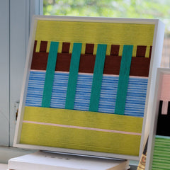Angled side view of the Woodland Mirror framed textile art resting on a surface, showcasing the dimensional depth of the woven green and blue yarn inside a white frame.