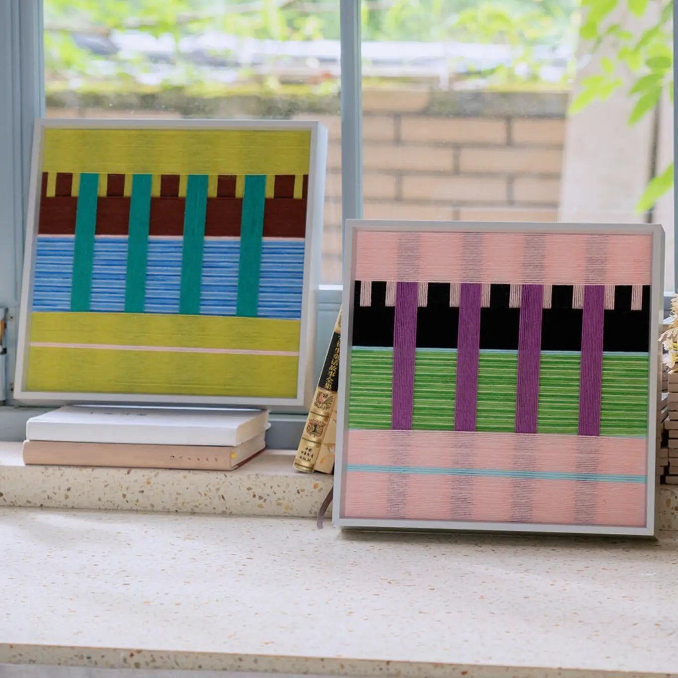 Green and blue woven textile art sitting on stacked books next to a pink variant on a low shelf near a bright window.